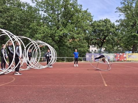 Gruppe von Sportlern trainiert auf einem roten Platz mit großen Reifen; eine Person rollt gerade durch einen Reifen.
