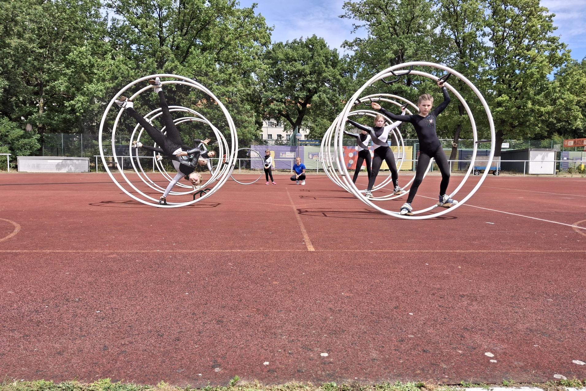 Kinder in Schwarz führen Tricks in großen, weißen Zirkusschwingen auf einem roten Sportplatz aus.
