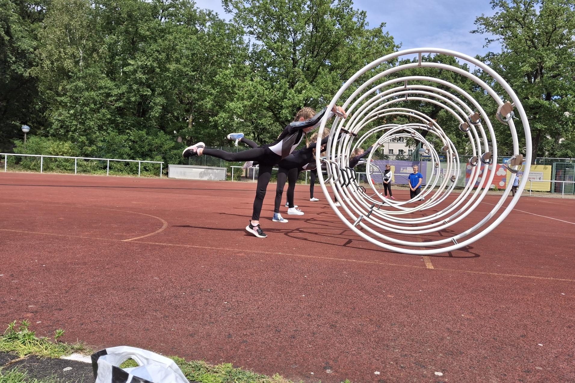 Sportler üben im Freien mit Trainingsgeräten auf einem roten Sportplatz, umgeben von grünen Bäumen und anderen Teilnehmern.