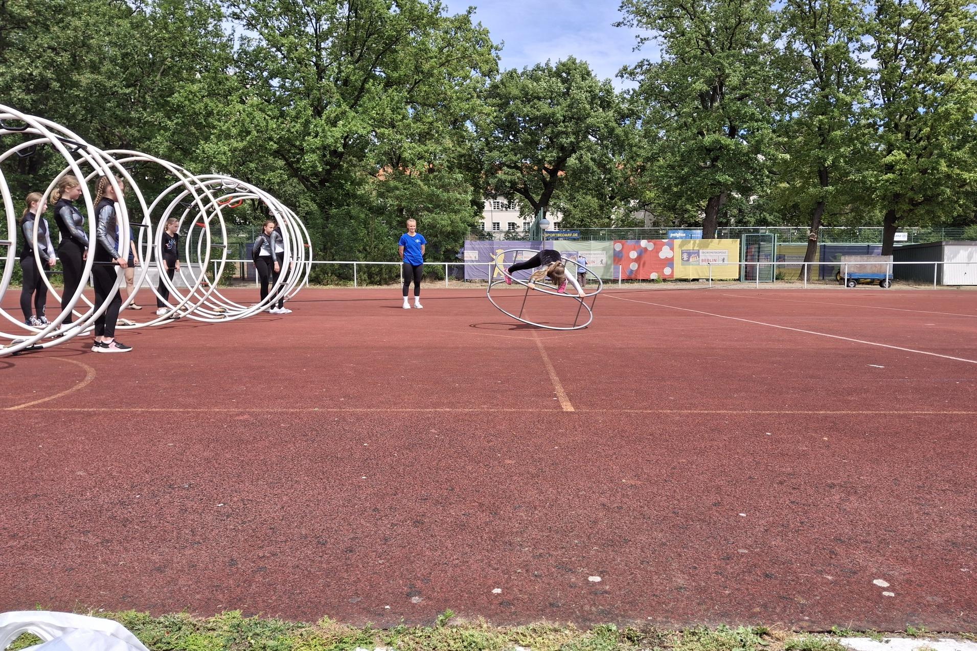 Gruppe von Sportlern trainiert auf einem roten Platz mit großen Reifen; eine Person rollt gerade durch einen Reifen.