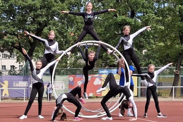 Gruppe von Mädchen in Sportanzügen bildet kunstvolle Pyramide auf einem Sportplatz mit einem großen Reifen im Hintergrund.