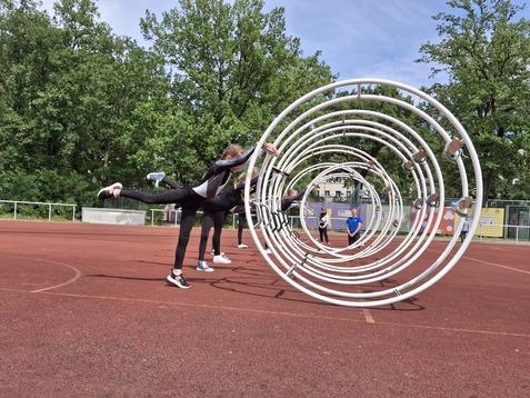 Sportler üben im Freien mit Trainingsgeräten auf einem roten Sportplatz, umgeben von grünen Bäumen und anderen Teilnehmern.