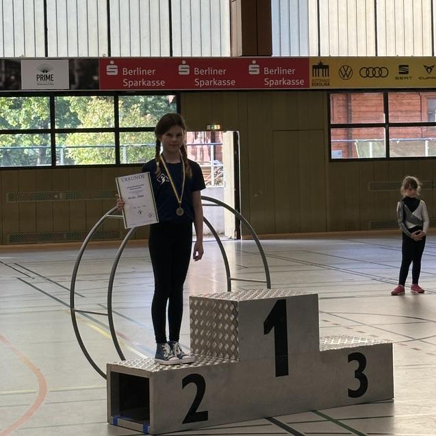 M&auml;dchen auf dem Siegerpodest h&auml;lt Urkunde und Medaille, im Hintergrund zwei weitere Teilnehmerinnen in Sportkleidung.
