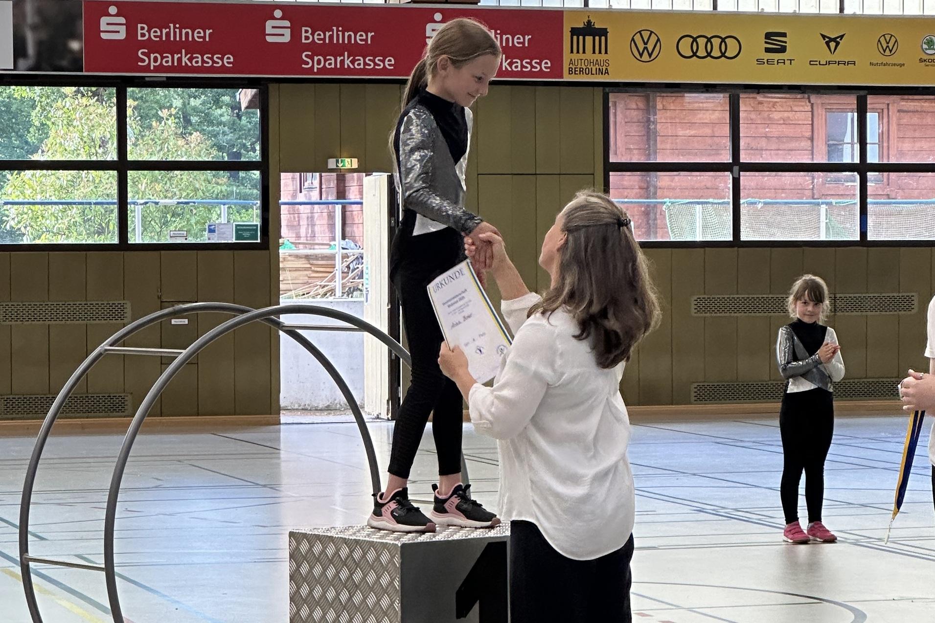 M&auml;dchen auf einem Siegertreppchen erh&auml;lt eine Urkunde von einer Frau in einer Sporthalle. Hintergrund mit weiteren Personen.