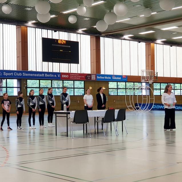 Szenenbild einer Sportveranstaltung mit M&auml;dchen in Sportanz&uuml;gen, einem Tisch und einer Wartenden im Hintergund.