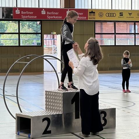 Mädchen auf einem Siegertreppchen erhält eine Urkunde von einer Frau in einer Sporthalle. Hintergrund mit weiteren Personen.