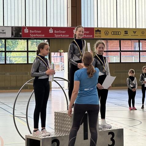 Drei Mädchen mit Medaillen stehen auf dem Podest, während eine Trainerin ihnen ihre Urkunden überreicht.