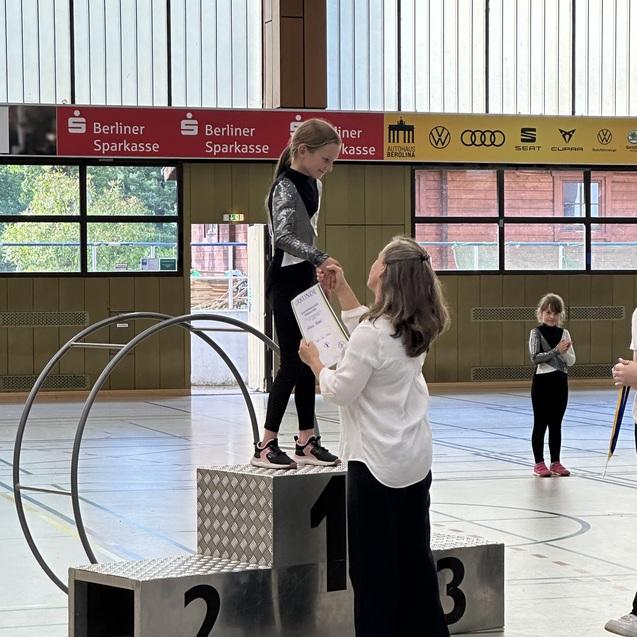 M&auml;dchen auf einem Siegertreppchen erh&auml;lt eine Urkunde von einer Frau in einer Sporthalle. Hintergrund mit weiteren Personen.