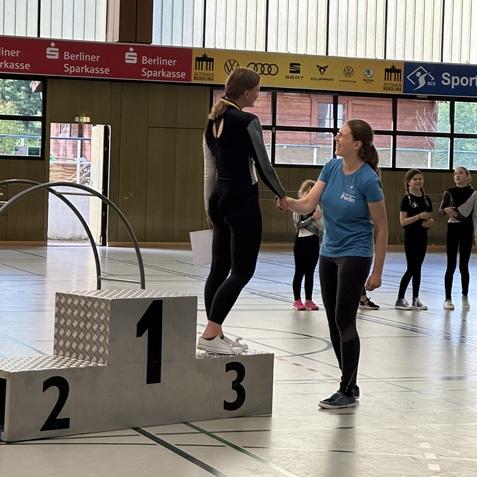 Zwei Sportlerinnen auf einem Siegerpodest, eine erh&auml;lt eine Medaille von einer Trainerin in einem Sporthallen-Setting.