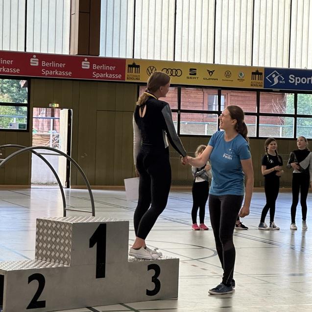 Zwei Sportlerinnen auf einem Siegerpodest, eine erh&auml;lt eine Medaille von einer Trainerin in einem Sporthallen-Setting.
