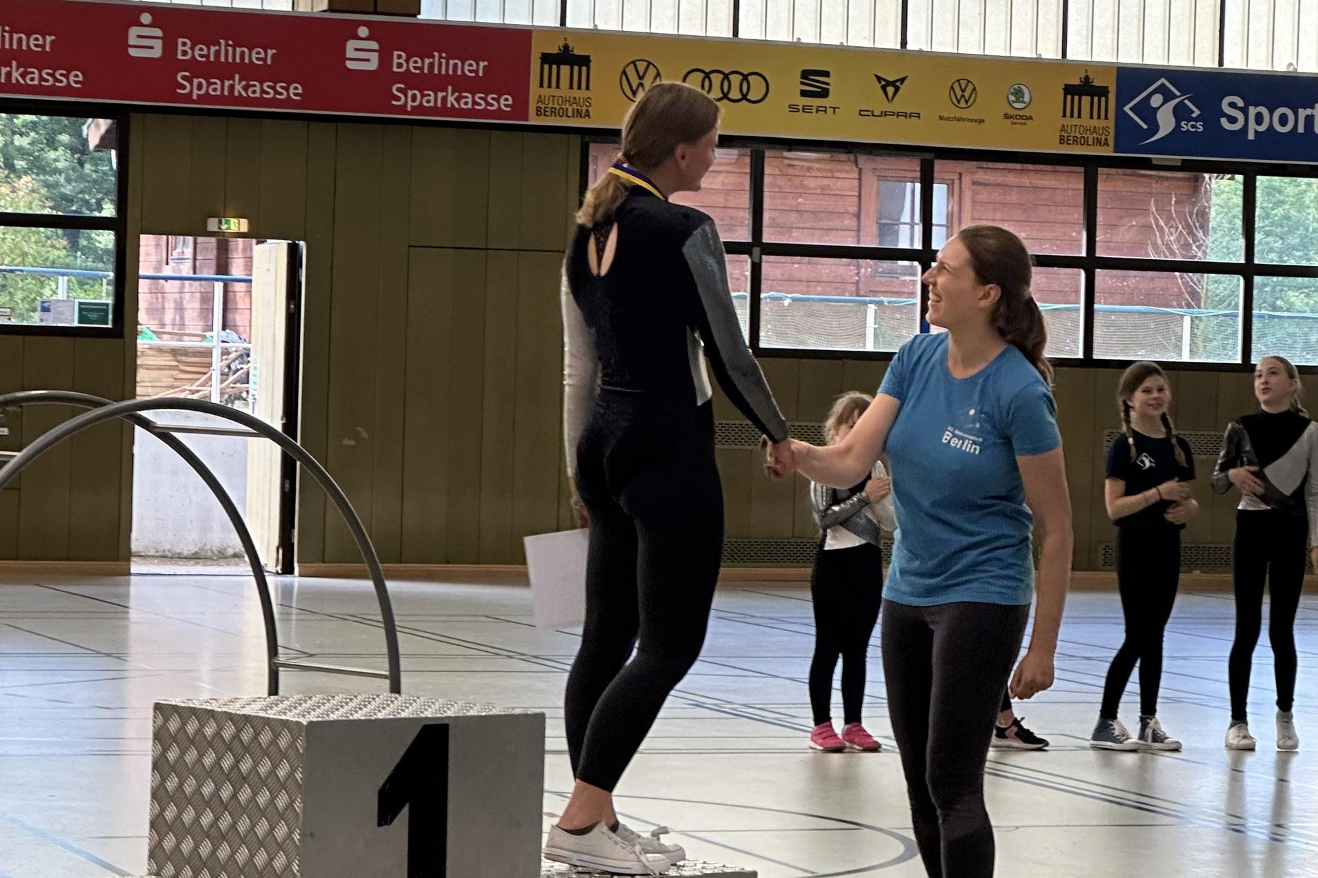 Zwei Sportlerinnen auf einem Siegerpodest, eine erh&auml;lt eine Medaille von einer Trainerin in einem Sporthallen-Setting.