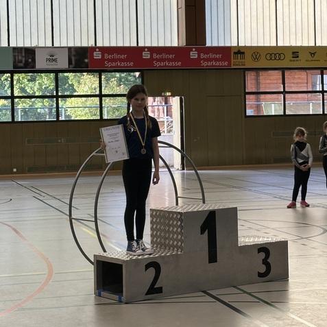 Mädchen auf dem Siegerpodest hält Urkunde und Medaille, im Hintergrund zwei weitere Teilnehmerinnen in Sportkleidung.