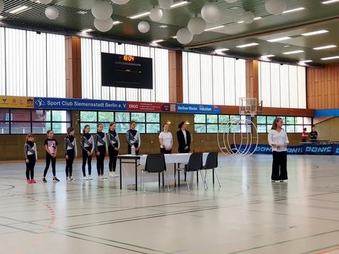 Szenenbild einer Sportveranstaltung mit M&auml;dchen in Sportanz&uuml;gen, einem Tisch und einer Wartenden im Hintergund.