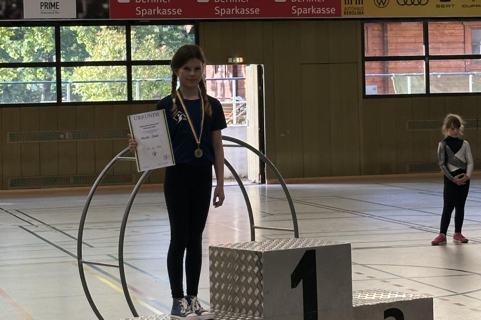 M&auml;dchen auf dem Siegerpodest h&auml;lt Urkunde und Medaille, im Hintergrund zwei weitere Teilnehmerinnen in Sportkleidung.