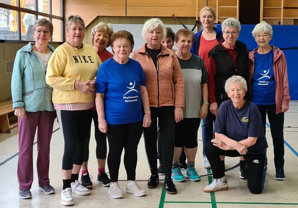 Gruppe älterer Frauen in Sportkleidung posiert gemeinsam in einer Turnhalle. Einige tragen T-Shirts mit Logo.