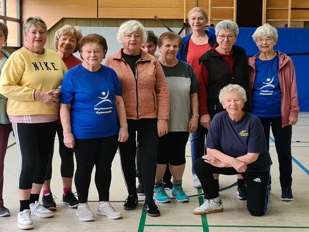Gruppe älterer Frauen in Sportkleidung posiert gemeinsam in einer Turnhalle. Einige tragen T-Shirts mit Logo.