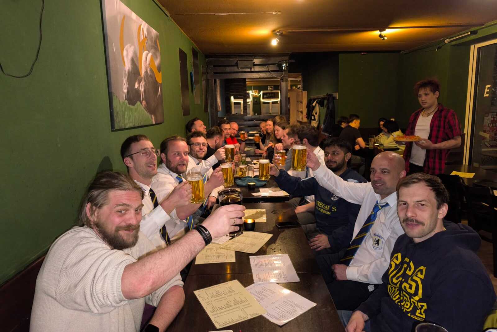 Gruppierung von Männern am Tisch in einer Bar, die mit Biergläsern anstoßen. Hintergrund mit grüner Wand und Bildern.