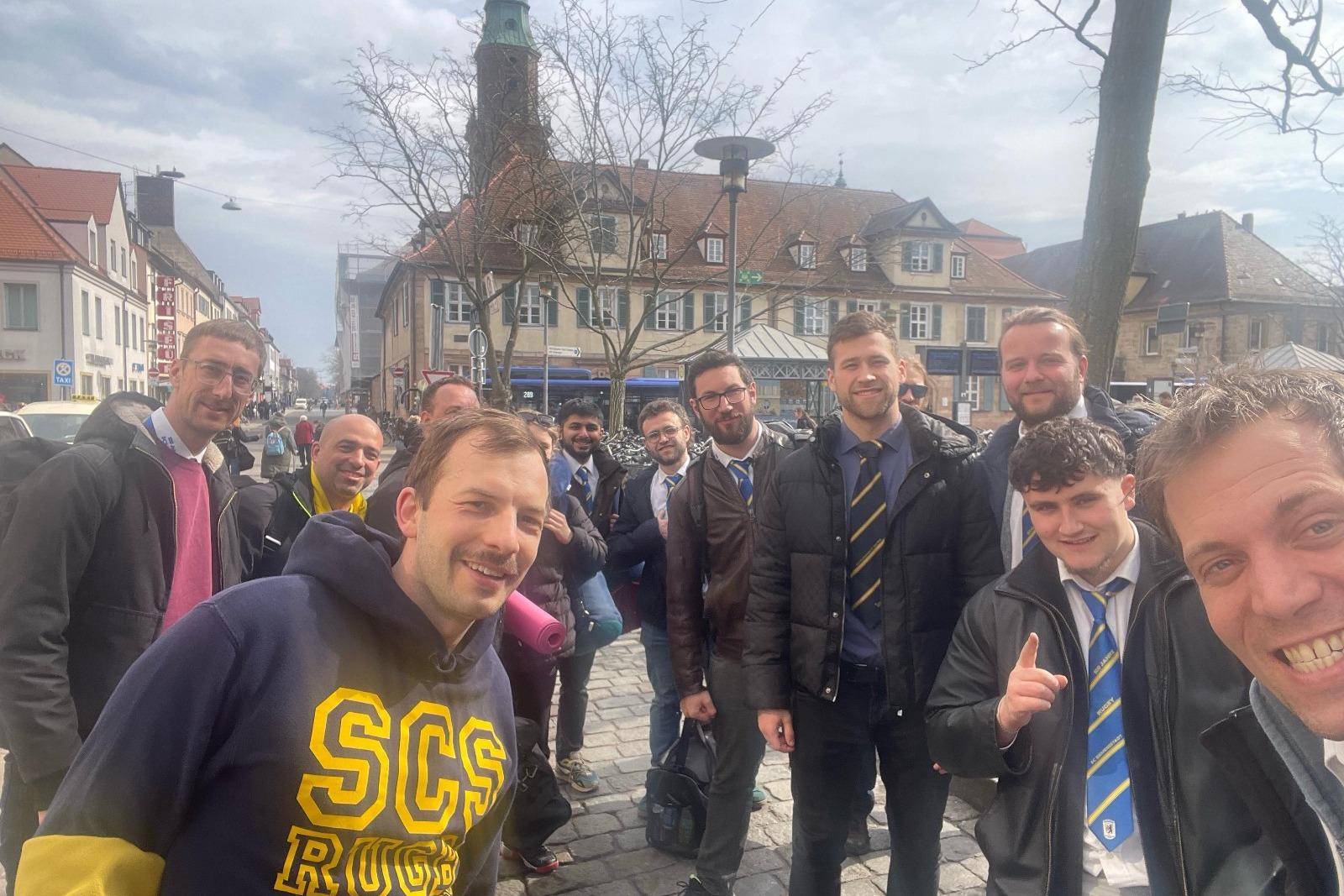 Gruppe von Männern posiert lächelnd auf einer Straße, historische Gebäude und Bäume im Hintergrund, bewölkter Himmel.