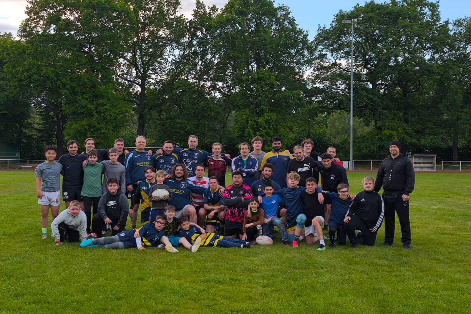 Gruppenfoto von Spielern und Trainern auf einem Rasenplatz nach dem Rugby-Training, umgeben von Bäumen.
