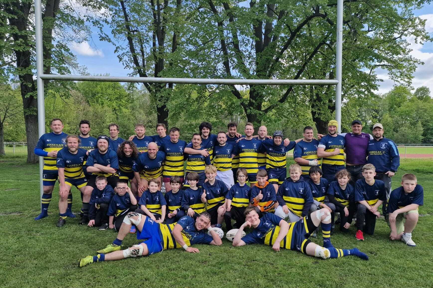 Rugby-Team mit Spielern verschiedenen Alters posiert lächelnd auf einem grünen Feld, umgeben von Bäumen und Torpfosten.