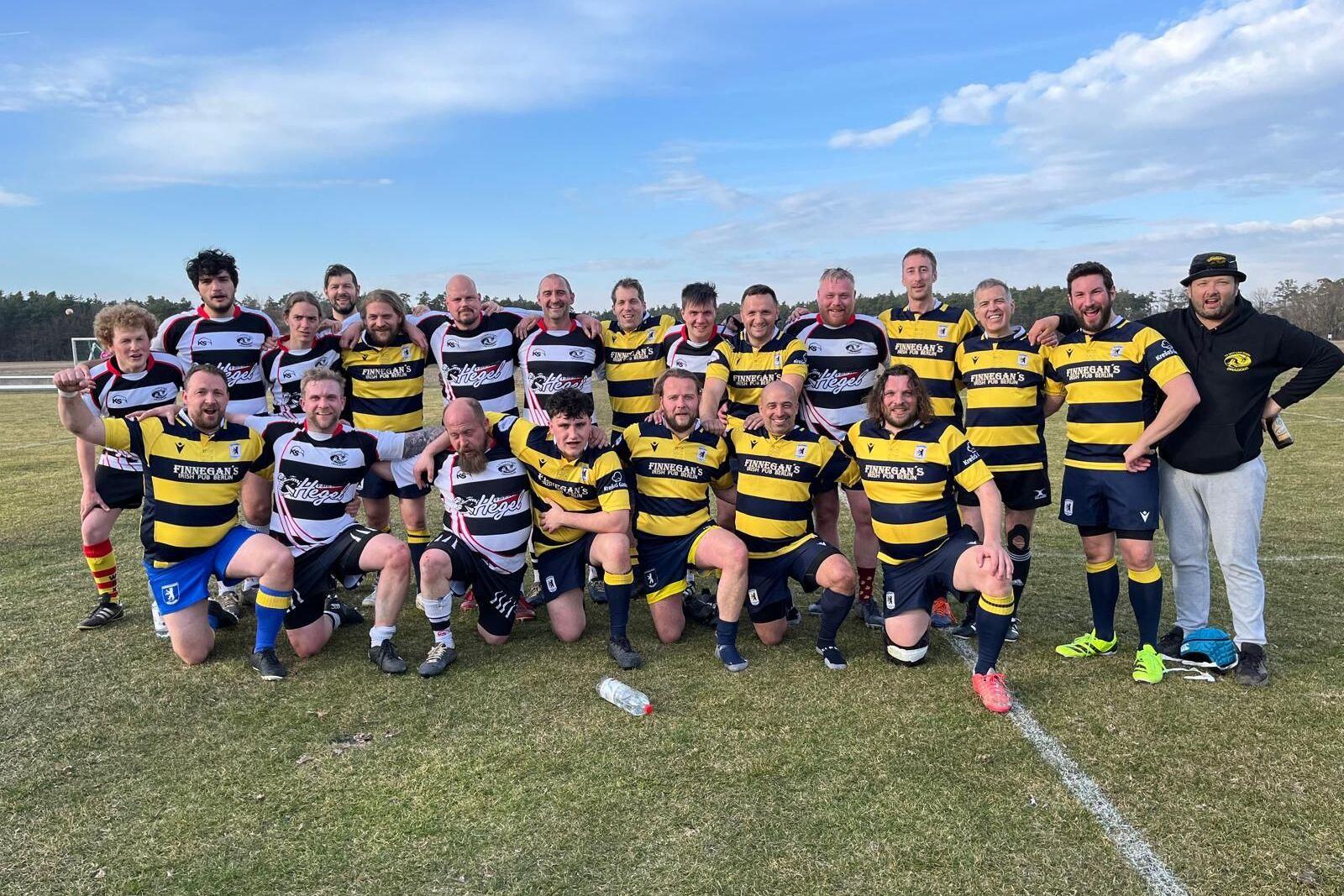 Gruppenfoto von Rugbyspielern in schwarz-gelben und rot-weißen Trikots auf einem Außenfeld, umgeben von Bäumen.