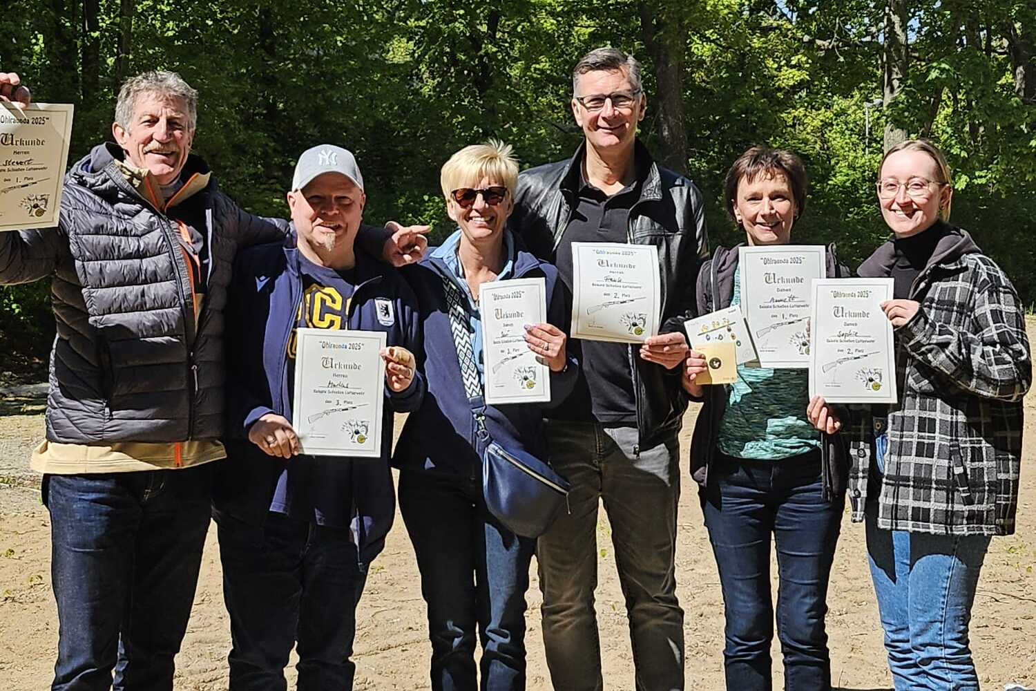 Sechs Personen stehen im Freien und halten Urkunden in den Händen, umgeben von grünen Bäumen und sandigem Boden.