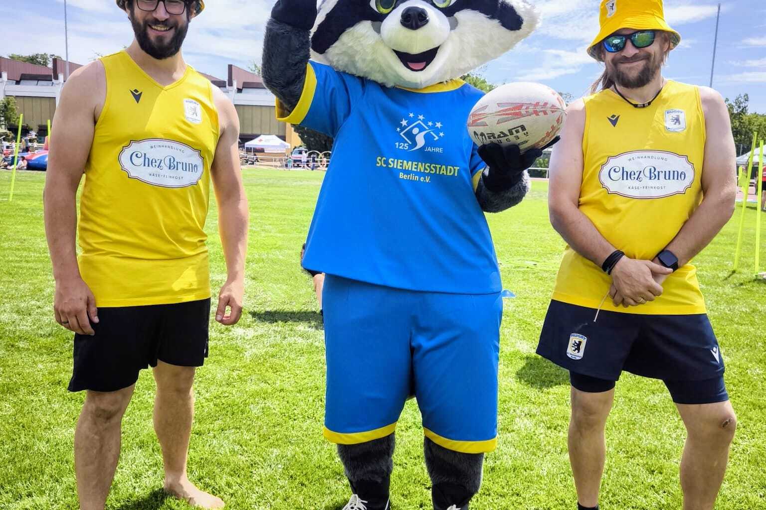 Drei Personen stehen auf einer grünen Wiese, zwei im Sportoutfit und ein Maskottchen mit Rugbyball in der Mitte.
