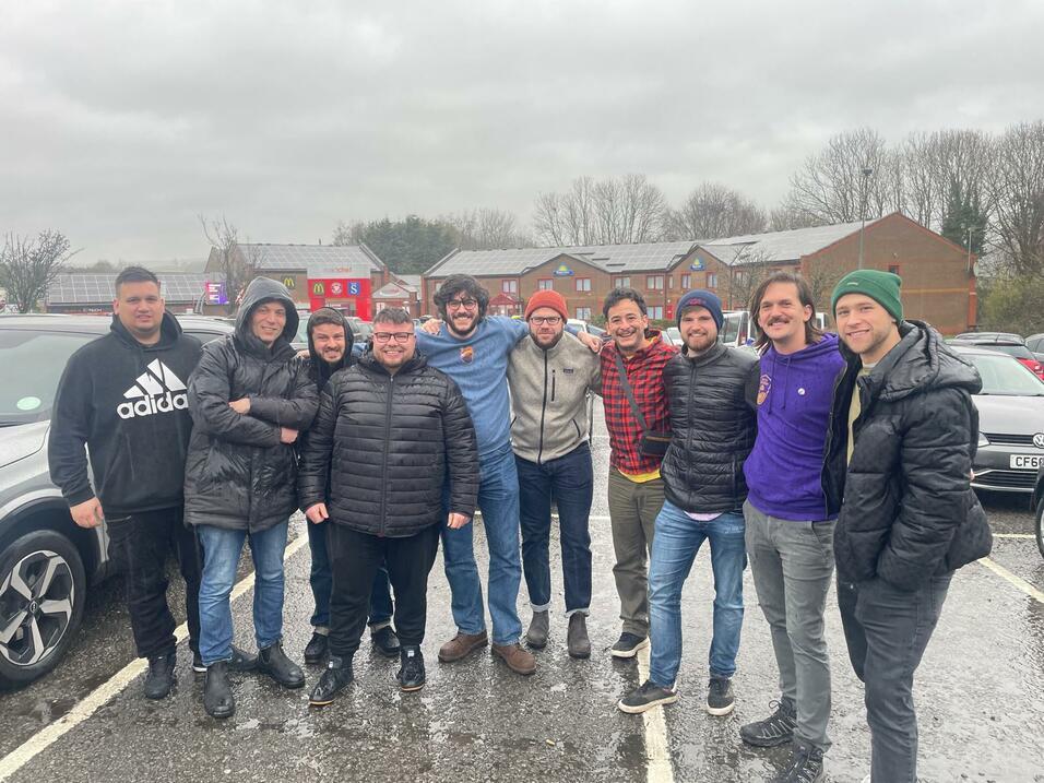 Gruppe von zehn Männern in wetterfester Kleidung lächelt gemeinsam auf einem Parkplatz bei bewölktem Himmel.