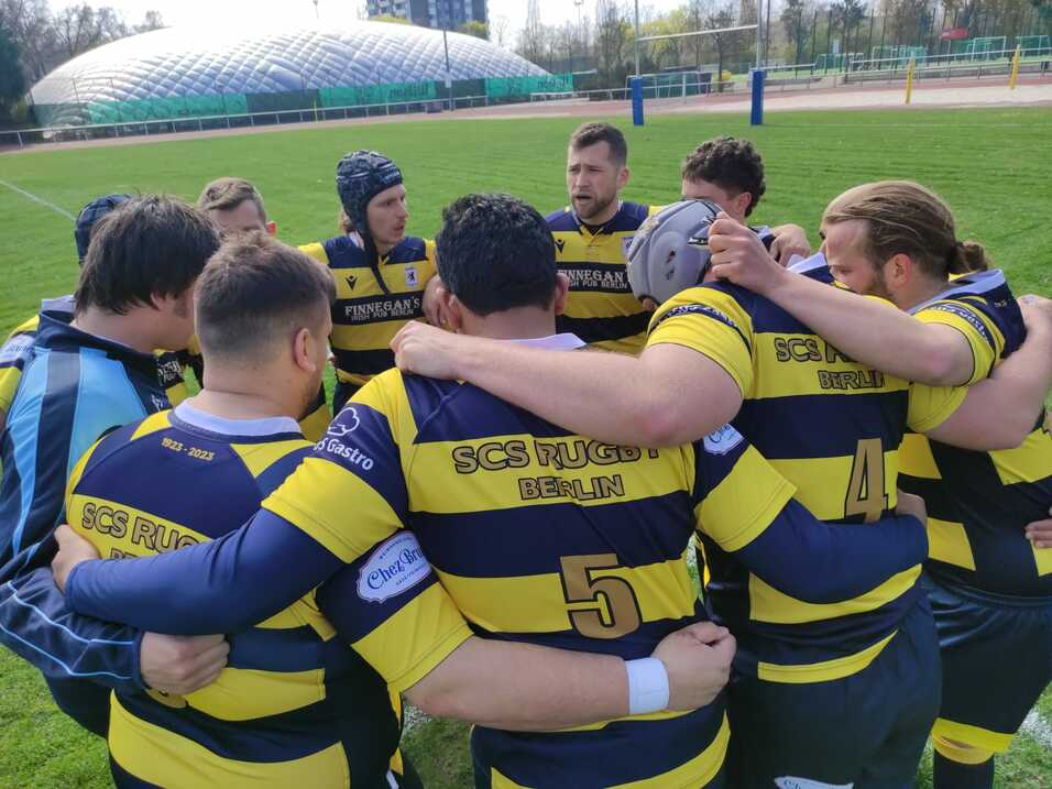 Eine Gruppe von Rugbyspielern in gestreiften Trikots bildet einen Kreis auf einem grünen Spielfeld.