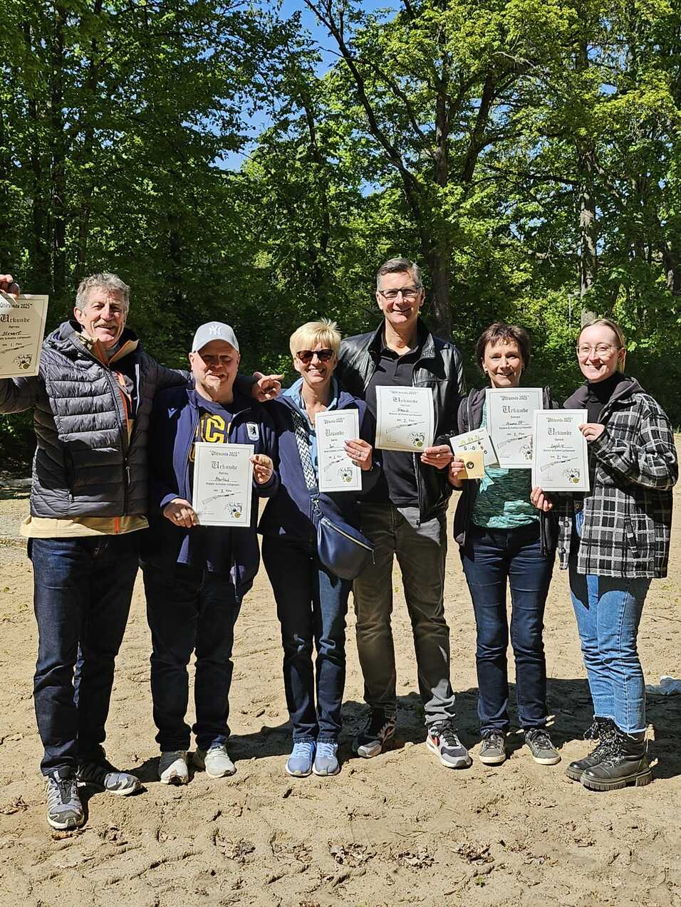 Sechs Personen stehen im Freien und halten Urkunden in den Händen, umgeben von grünen Bäumen und sandigem Boden.