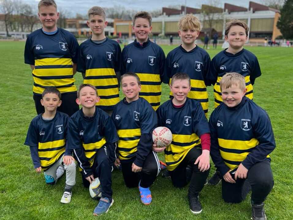 Gruppe von zehn Kindern in Rugby-Trikots mit gelben und blauen Streifen, l&auml;cheln auf einem Spielfeld.
