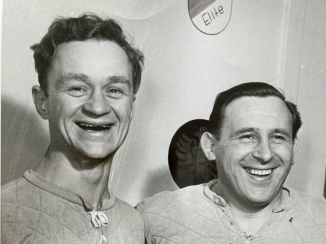 Zwei lächelnde Männer in sportlichen Trikots mit dem Buchstaben "S" stehen nebeneinander vor einem hellen Hintergrund.