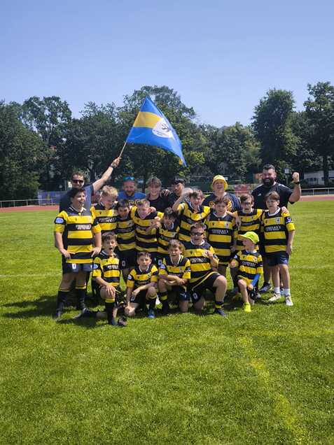 Gruppenshot von Kindern in gelb-schwarzen Trikots mit einem Flagge, vor einer Wiese und blauem Himmel.