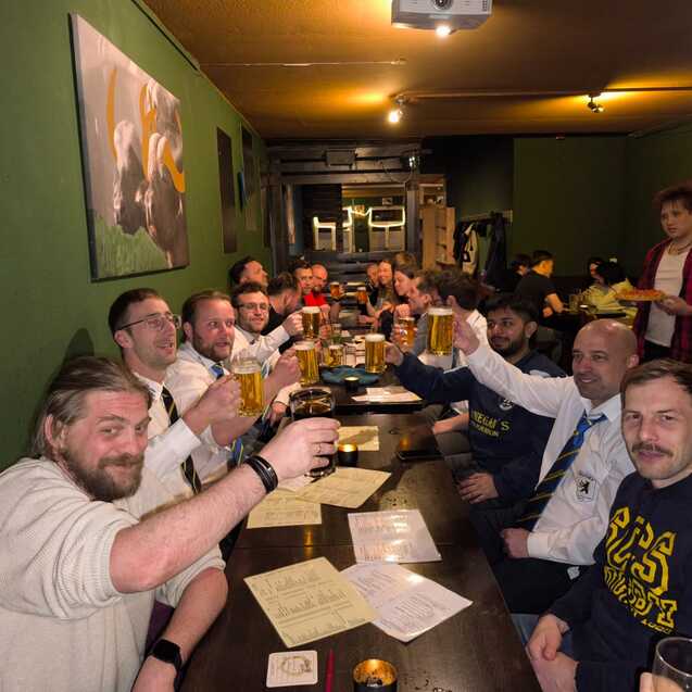 Gruppierung von Männern am Tisch in einer Bar, die mit Biergläsern anstoßen. Hintergrund mit grüner Wand und Bildern.
