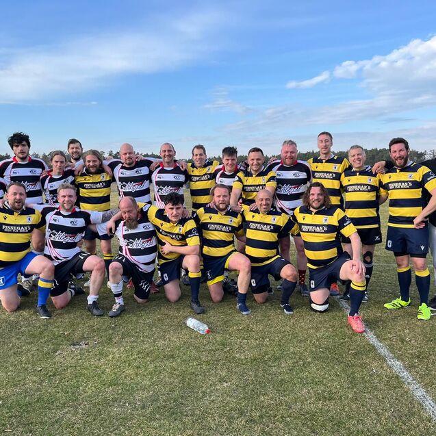 Gruppenfoto von zwei Rugbyteams in einheitlicher Kleidung auf einem Sportfeld, umgeben von Natur und blauem Himmel.