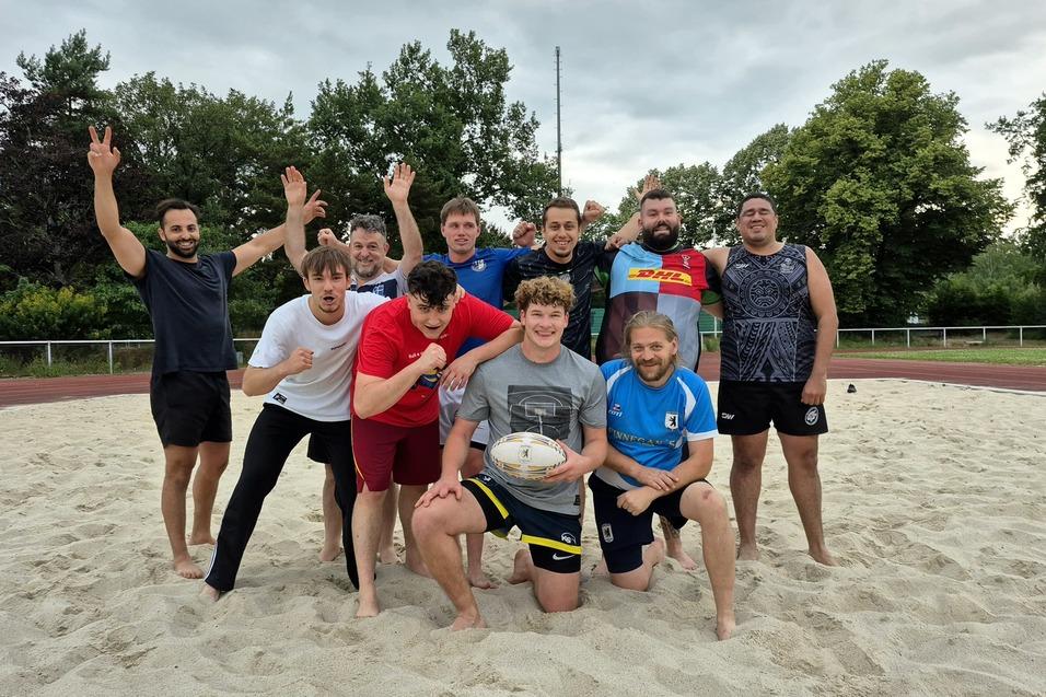 Gruppenshot von zehn Rugbyspielern auf einem Sandfeld, einige posieren, w&auml;hrend andere jubeln.