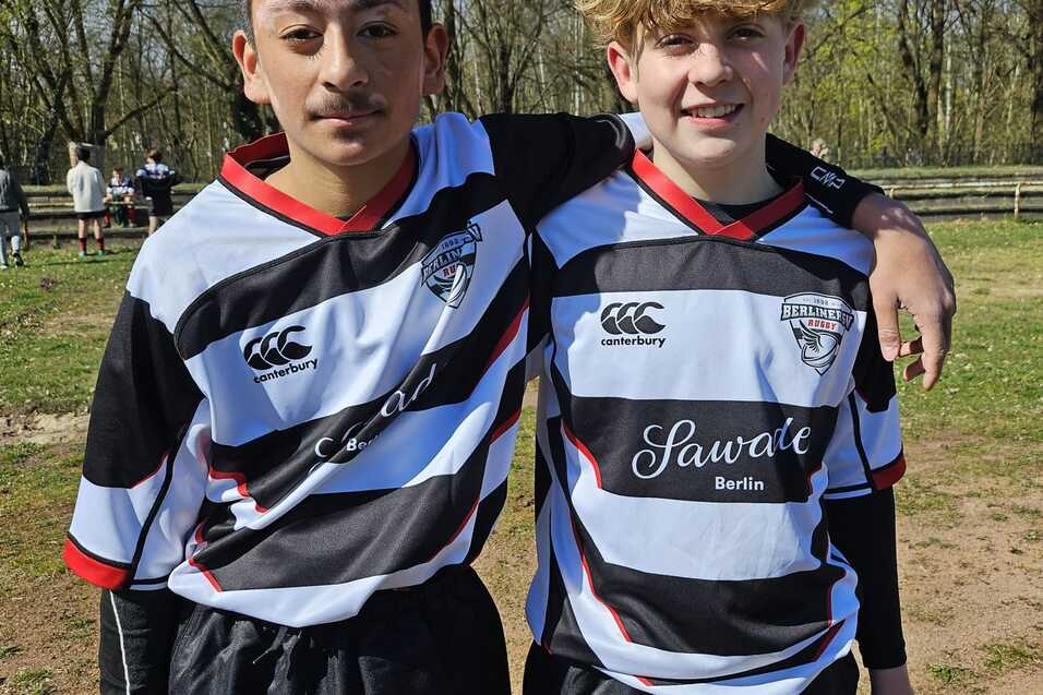 Zwei junge Fu&szlig;ballspieler stehen nebeneinander, tragen schwarze und wei&szlig;e Trikots und l&auml;cheln in die Kamera.