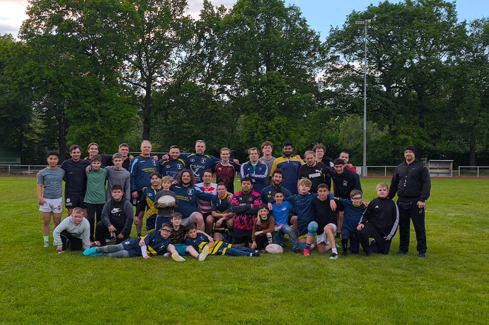 Gruppenfoto von Spielern und Trainern auf einem Rasenplatz nach dem Rugby-Training, umgeben von B&auml;umen.
