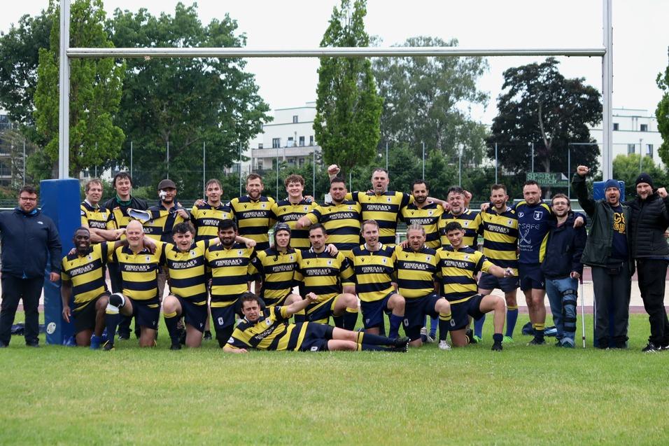 Gruppenfoto einer Rugby-Mannschaft in gestreiften Trikots, l&auml;chelnd auf einem Rasenplatz unter freiem Himmel.