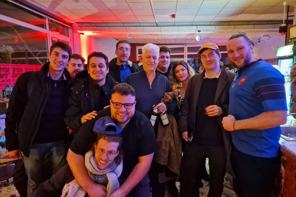 Gruppe von zehn Personen in einem Innenraum, teilweise lachend und mit Getr&auml;nken, vor einer bunten Hintergrundbeleuchtung.