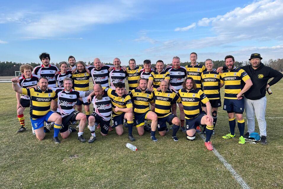 Gruppenfoto von Rugbyspielern in schwarz-gelben und rot-wei&szlig;en Trikots auf einem Au&szlig;enfeld, umgeben von B&auml;umen.
