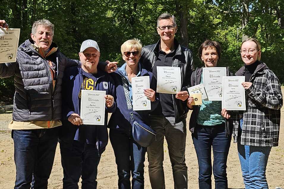 Sechs Personen stehen im Freien und halten Urkunden in den H&auml;nden, umgeben von gr&uuml;nen B&auml;umen und sandigem Boden.