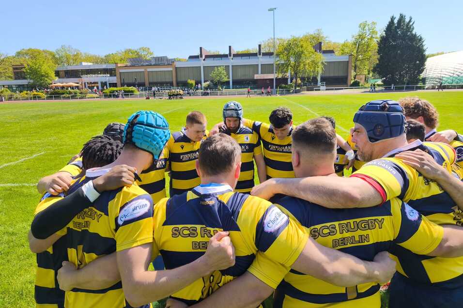 Rugby-Team in gelb-blauen Trikots bildet eine huddle auf einem gr&uuml;nen Platz unter blauem Himmel.