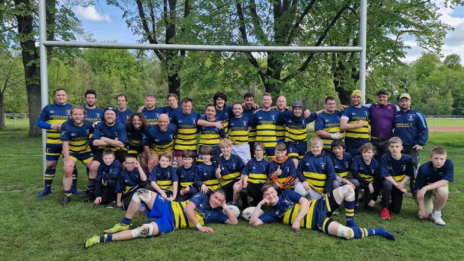 Rugby-Team mit Spielern verschiedenen Alters posiert lächelnd auf einem grünen Feld, umgeben von Bäumen und Torpfosten.