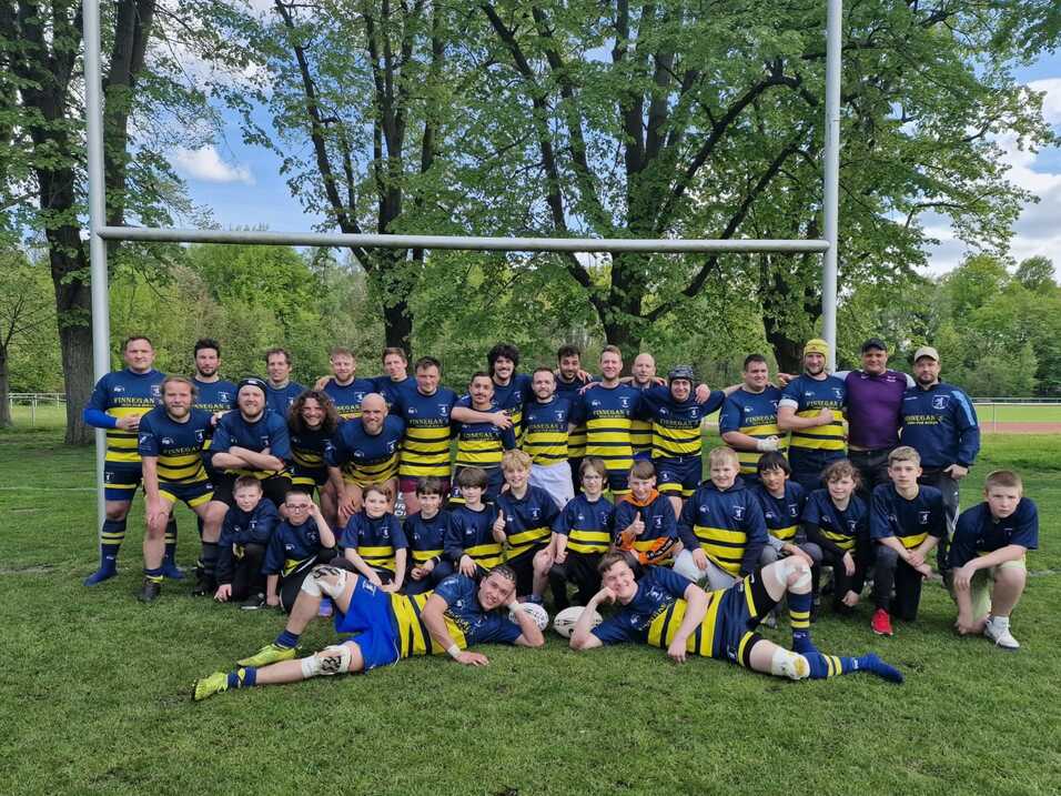 Rugby-Team mit Spielern verschiedenen Alters posiert lächelnd auf einem grünen Feld, umgeben von Bäumen und Torpfosten.