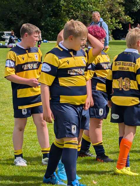 Vier Kinder in Rugbytrikots mit gelb-schwarzen Streifen stehen auf einem grünen Rasenplatz.