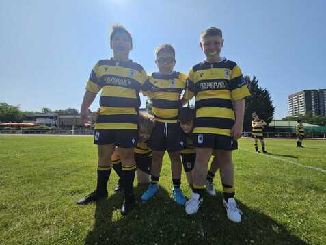 Vier Kinder in gestreiften Rugbytrikots stehen fröhlich auf einem Sportplatz. Sonniger Tag mit grünem Rasen im Hintergrund.