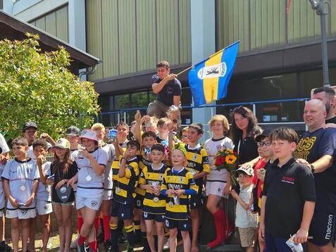 Gruppenfoto von Kindern in Rugby-Trikots mit Medaillen, einer hält eine Flagge, Erwachsene und ein Blumenstrauß sind ebenfalls zu sehen.
