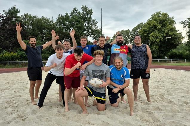 Gruppenshot von zehn Rugbyspielern auf einem Sandfeld, einige posieren, während andere jubeln.