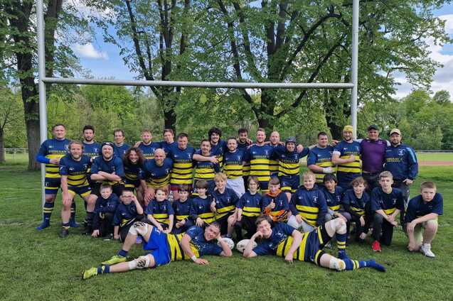 Rugby-Team mit Spielern verschiedenen Alters posiert l&auml;chelnd auf einem gr&uuml;nen Feld, umgeben von B&auml;umen und Torpfosten.